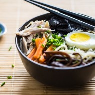 Zupa Ramen w miseczce z jajkiem, makaronem, marchewką, mięsem, grzybami i glonami. Na miseczce leżą pałeczki